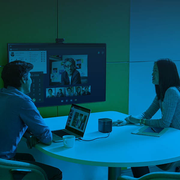 Two people in a small meeting room with a third person teleconferencing